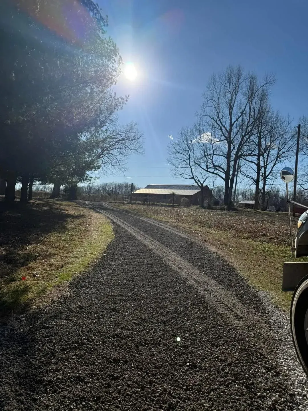 Sunlit gravel driveway curving past trees toward a small barn under a clear blue sky