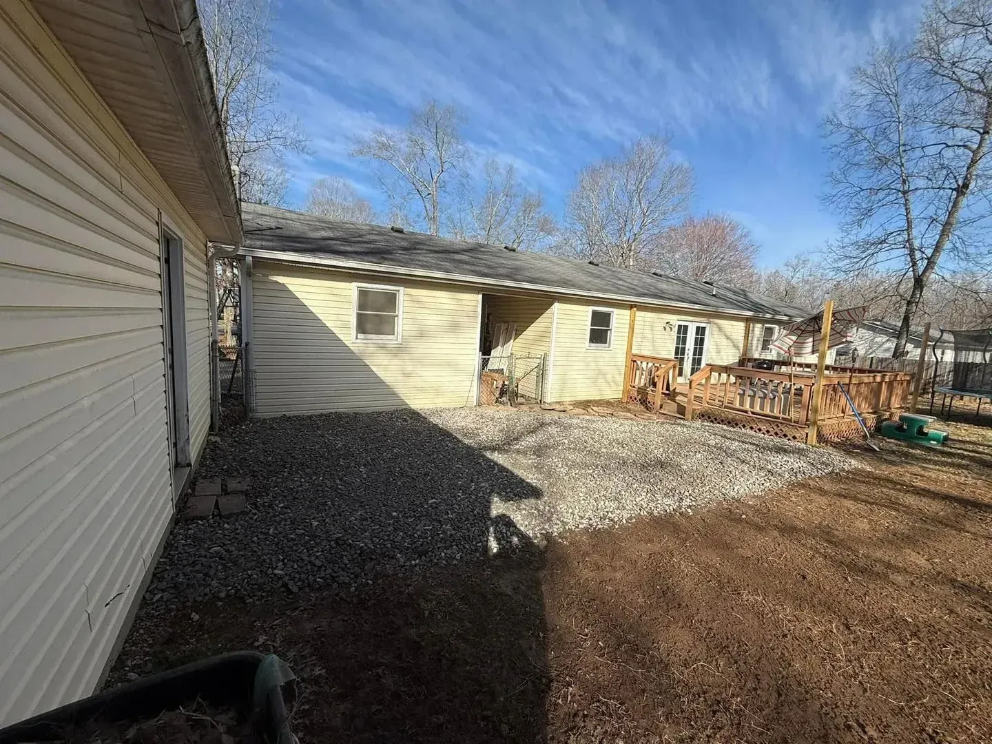 Backyard with beige house siding, gravel ground, and leafless trees under a clear sky