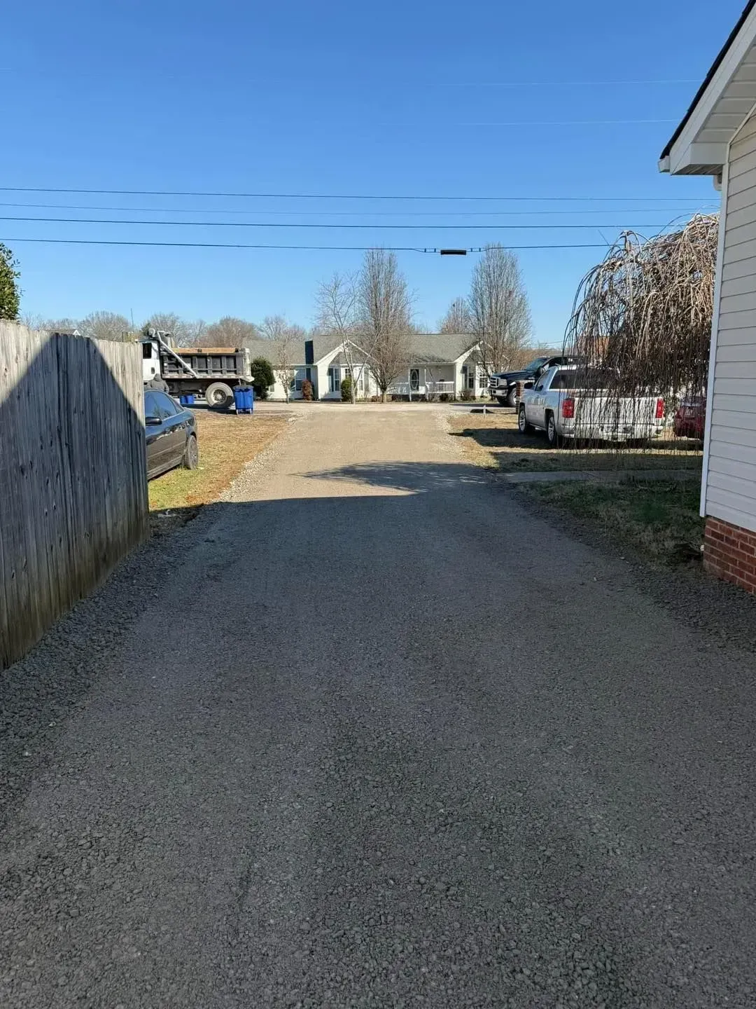 Gravel driveway leading to a rural yard with parked vehicles, a white house, and blue sky.