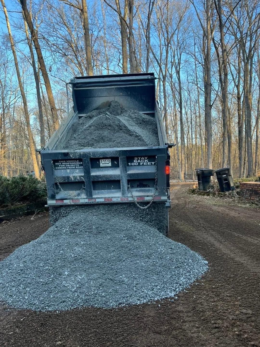 Dump truck unloading gray gravel on a dirt road in a wooded area.