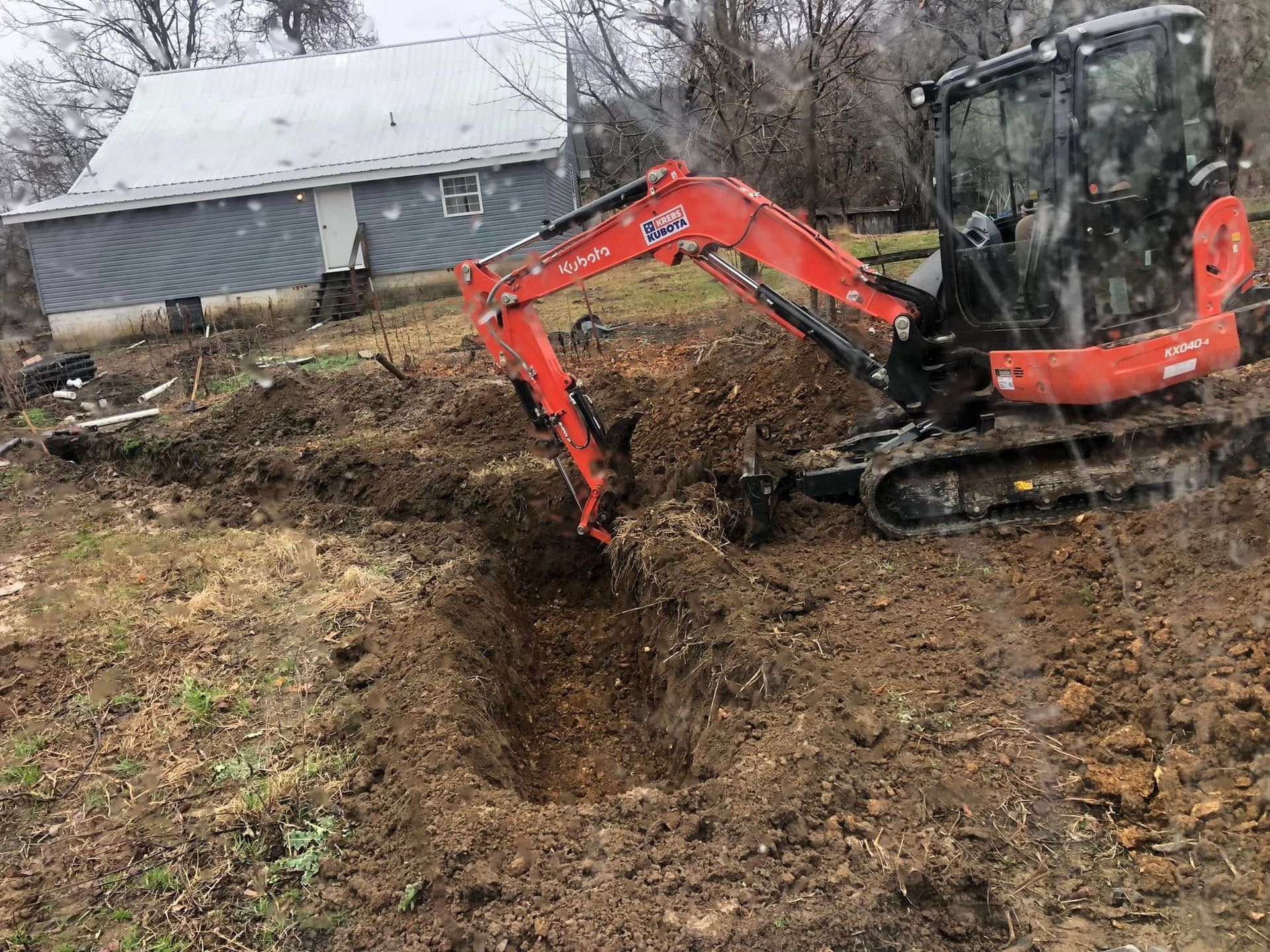 Red excavator digging a muddy ditch beside a house on a hillside