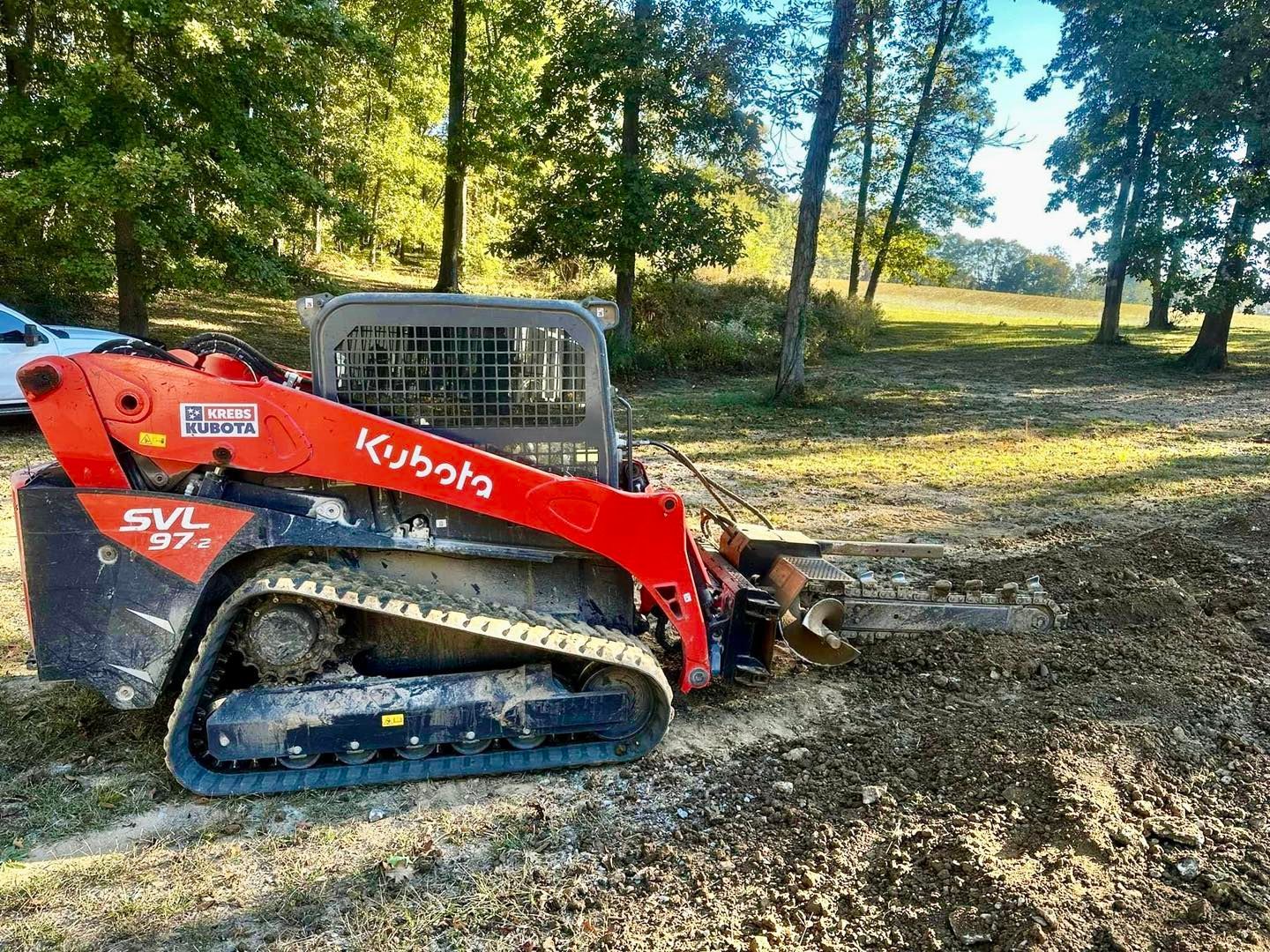 Red Kubota tracked loader on a dirt path in a wooded area, with a bucket attachment in front.