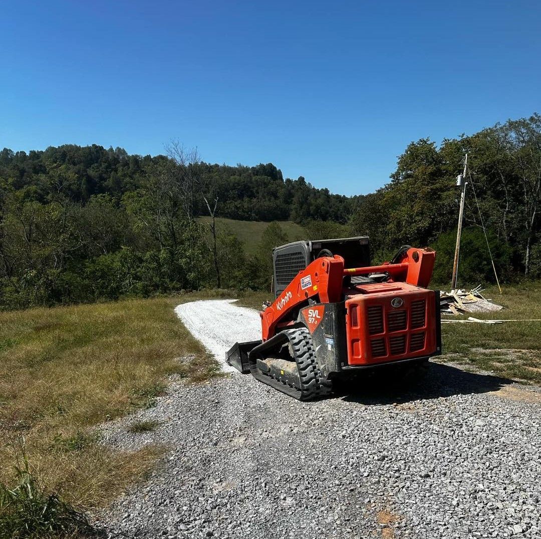Red excavator on a gravel road beside grassy hills under a clear blue sky