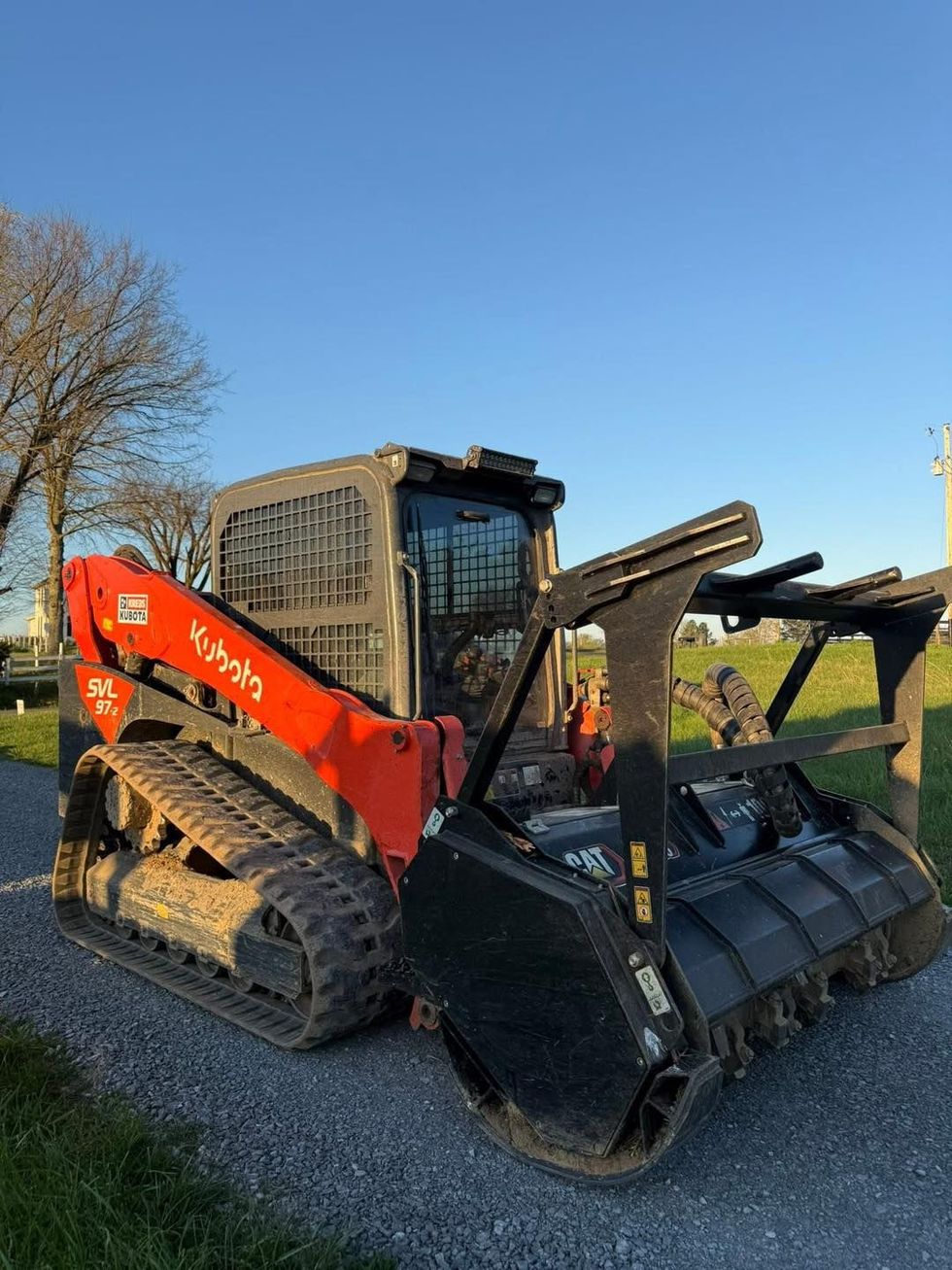 Orange Kubota compact track loader with attachments parked on grass under a blue sky
