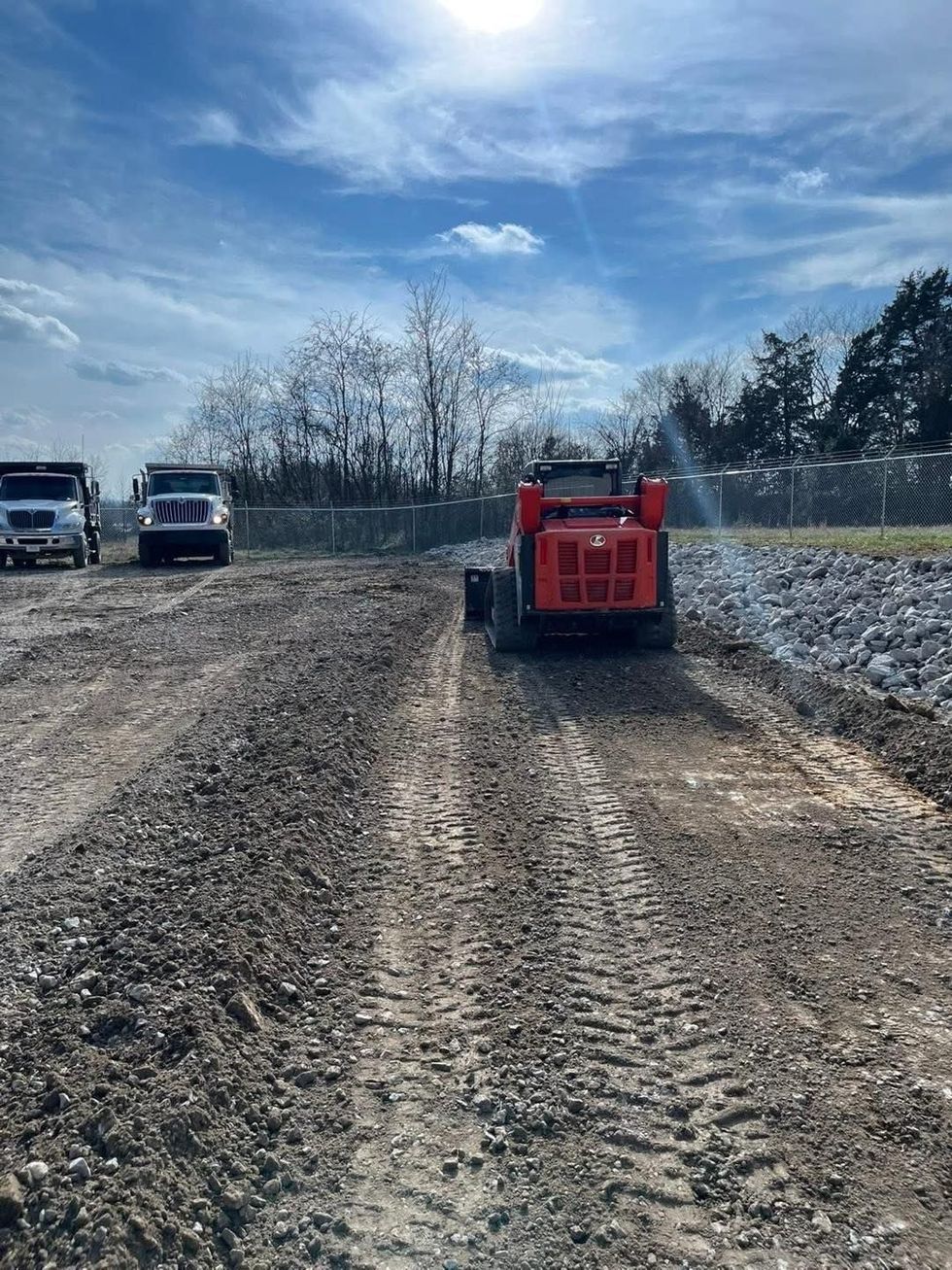 Red utility vehicle on a muddy dirt road under a bright blue sky, with trucks parked to the left.