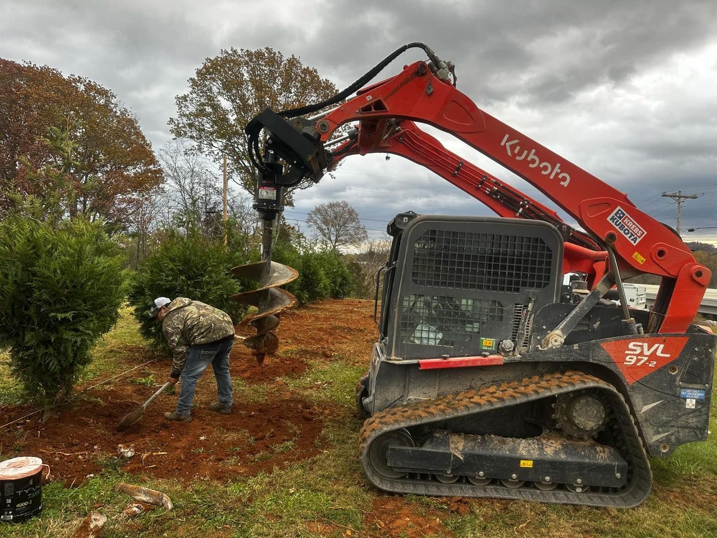 Red Kubota mini excavator clearing brush beside a worker in a wooded area