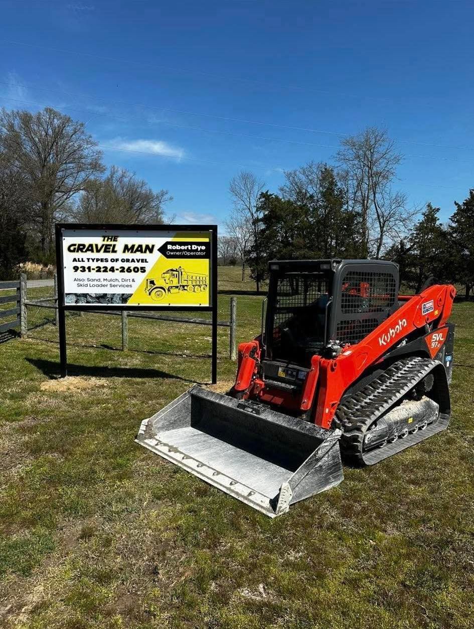 Red mini skid steer with bucket beside a gravel yard sign under a blue sky