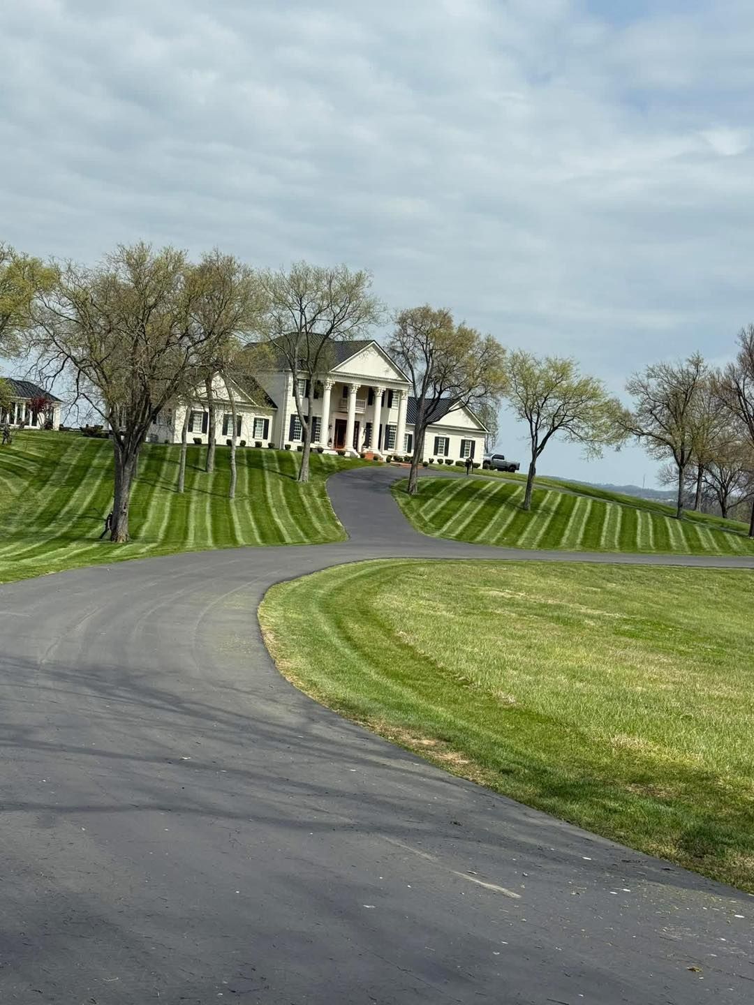 Grand white mansion on a grassy hill with a winding driveway and overcast sky
