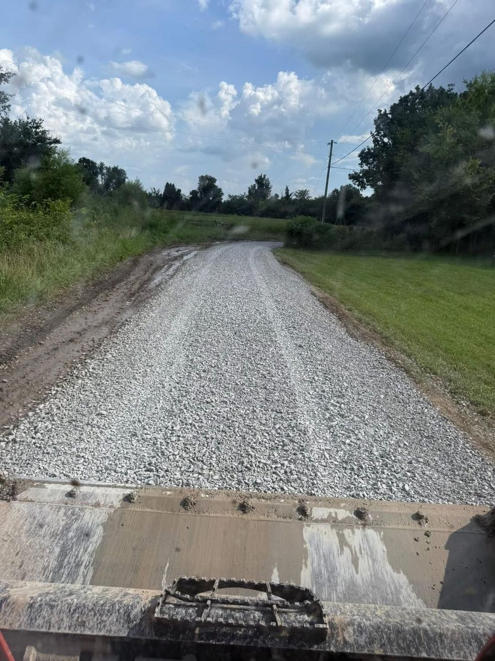 Gravel road ahead from a vehicle, bordered by grass and trees under a cloudy sky