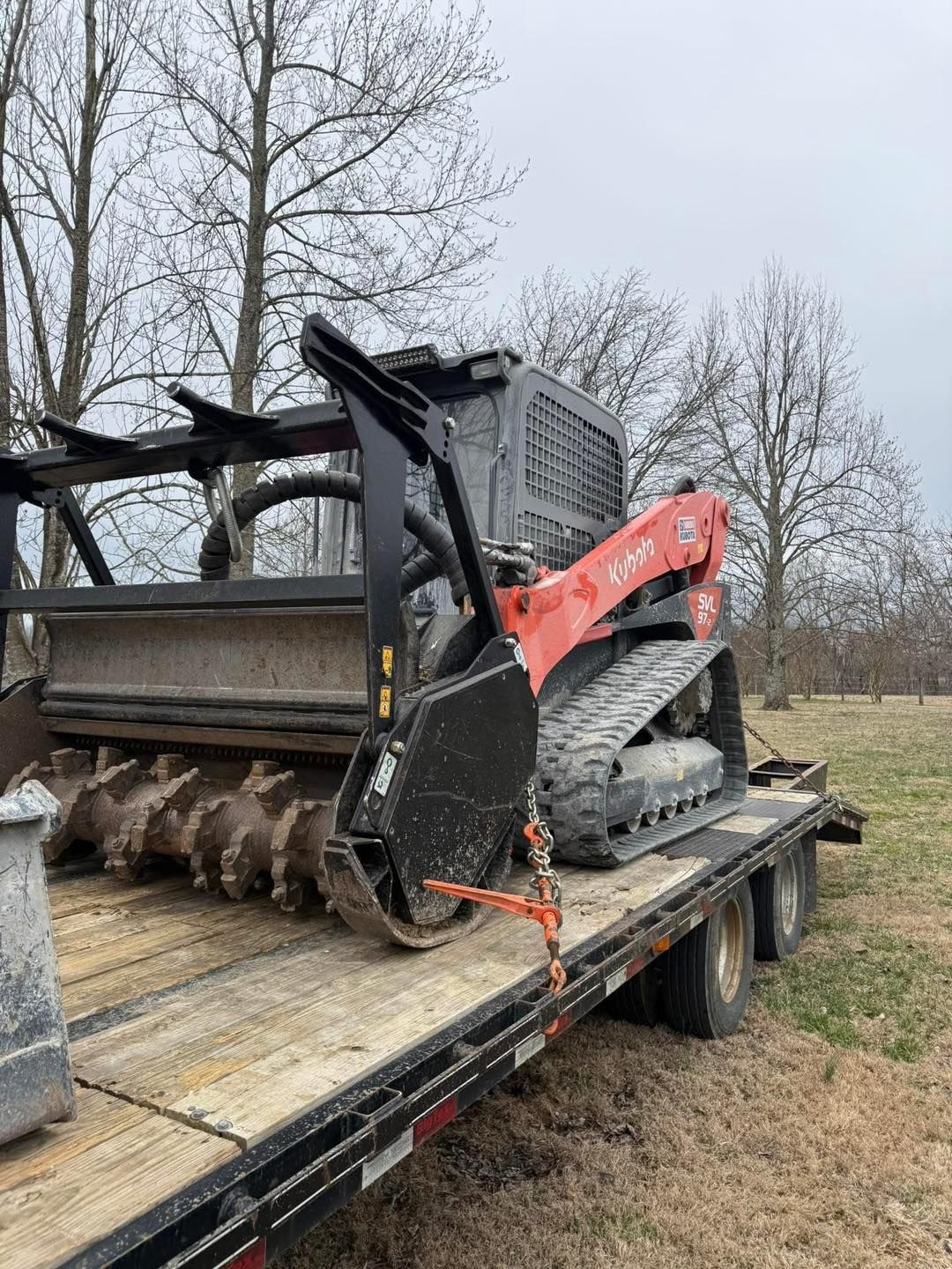 Small black skid steer with red arm on a flatbed trailer in a grassy yard, with trees in the background