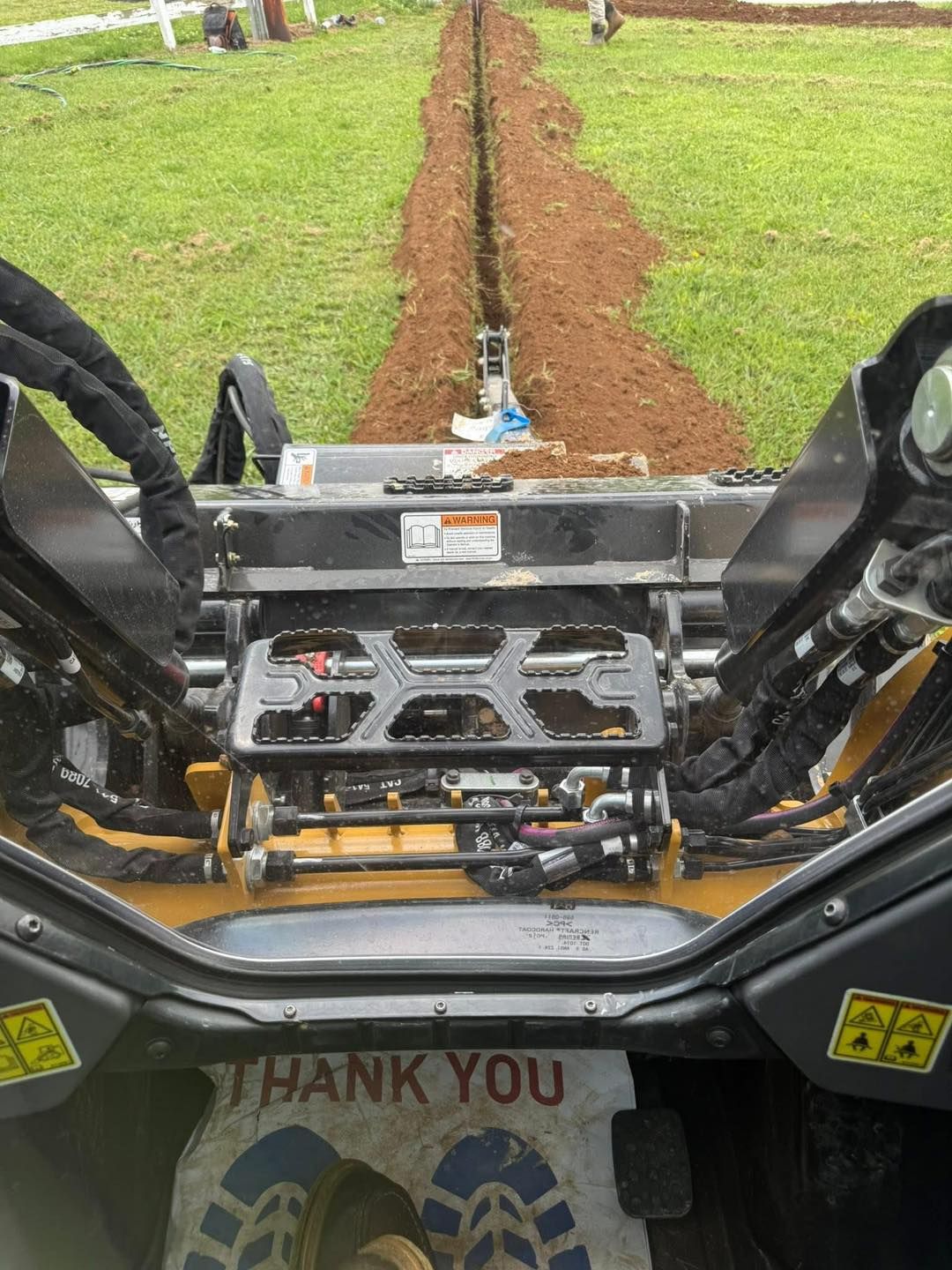 View from a machine cab digging a narrow trench through grassy ground