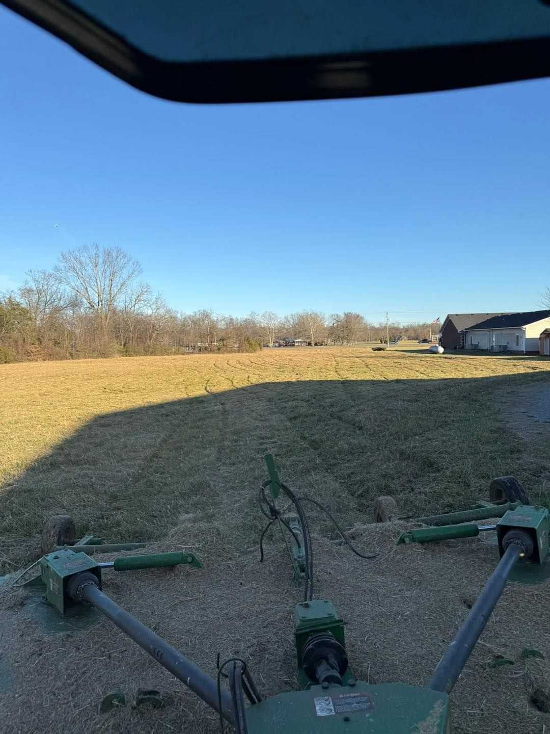 View from inside a mower over a grassy field with trees and houses in the distance.