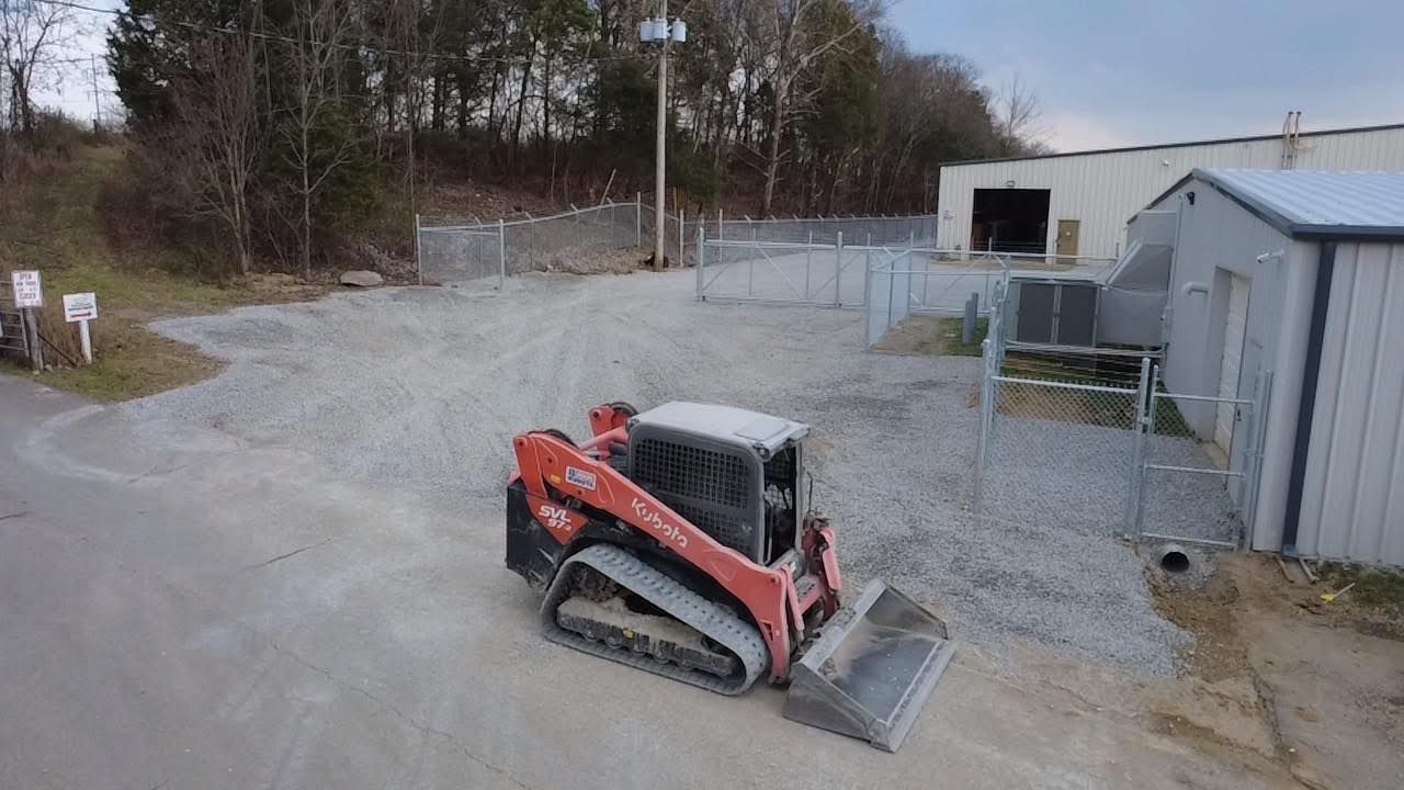 Red skid-steer loader with bucket on gravel in a fenced industrial yard near small buildings.