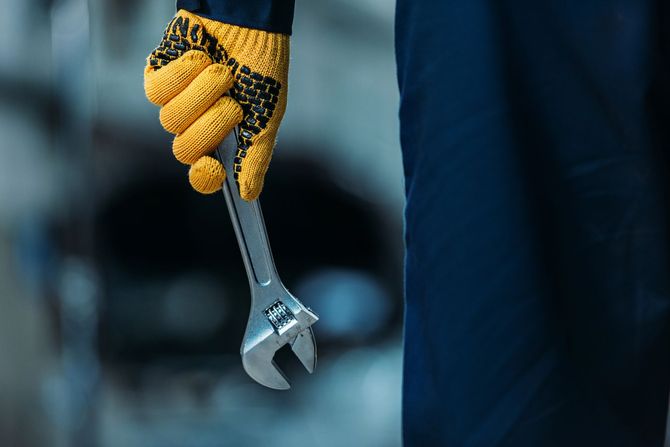 Mano guantata che impugna una chiave inglese argentata su uno sfondo industriale blu sfocato.