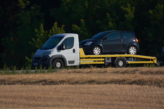 Un carro attrezzi bianco con pianale che trasporta un'auto hatchback scura ai margini di un campo, vicino agli alberi.