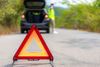 Triangolo di segnalazione rosso sulla strada vicino a un'auto in panne con il bagagliaio aperto e persona sfocata con giubbotto di sicurezza.