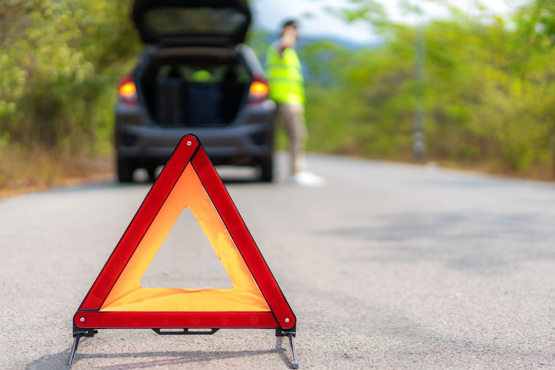 Un triangolo rosso di segnalazione su strada, accanto a un'auto con il bagagliaio aperto e una persona sfocata con indosso un giubbotto di sicurezza.