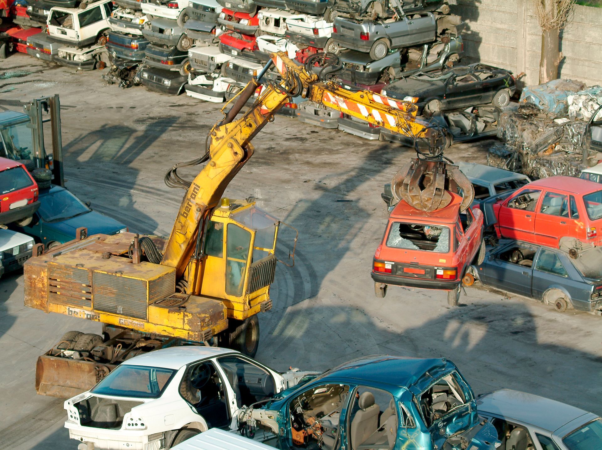 Un escavatore giallo sposta auto incidentate in una discarica, con cumuli di veicoli schiacciati tutt'intorno.