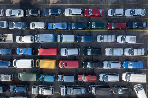 Veduta aerea di auto parcheggiate disposte in file ravvicinate in un parcheggio, tra cui veicoli rossi, blu, bianchi e gialli.