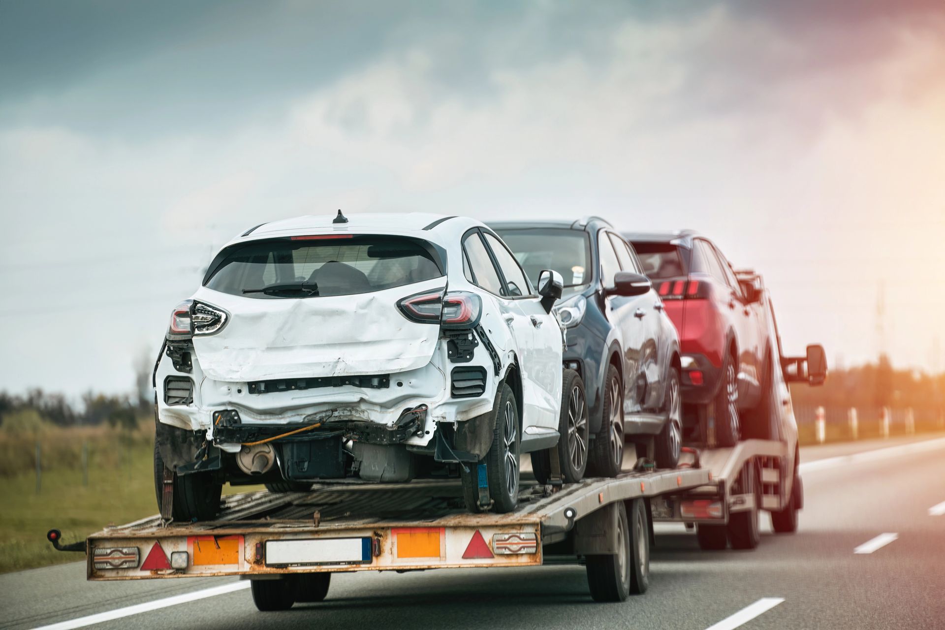 Un'auto bianca danneggiata su un rimorchio sta percorrendo un'autostrada, con altri veicoli alle sue spalle.