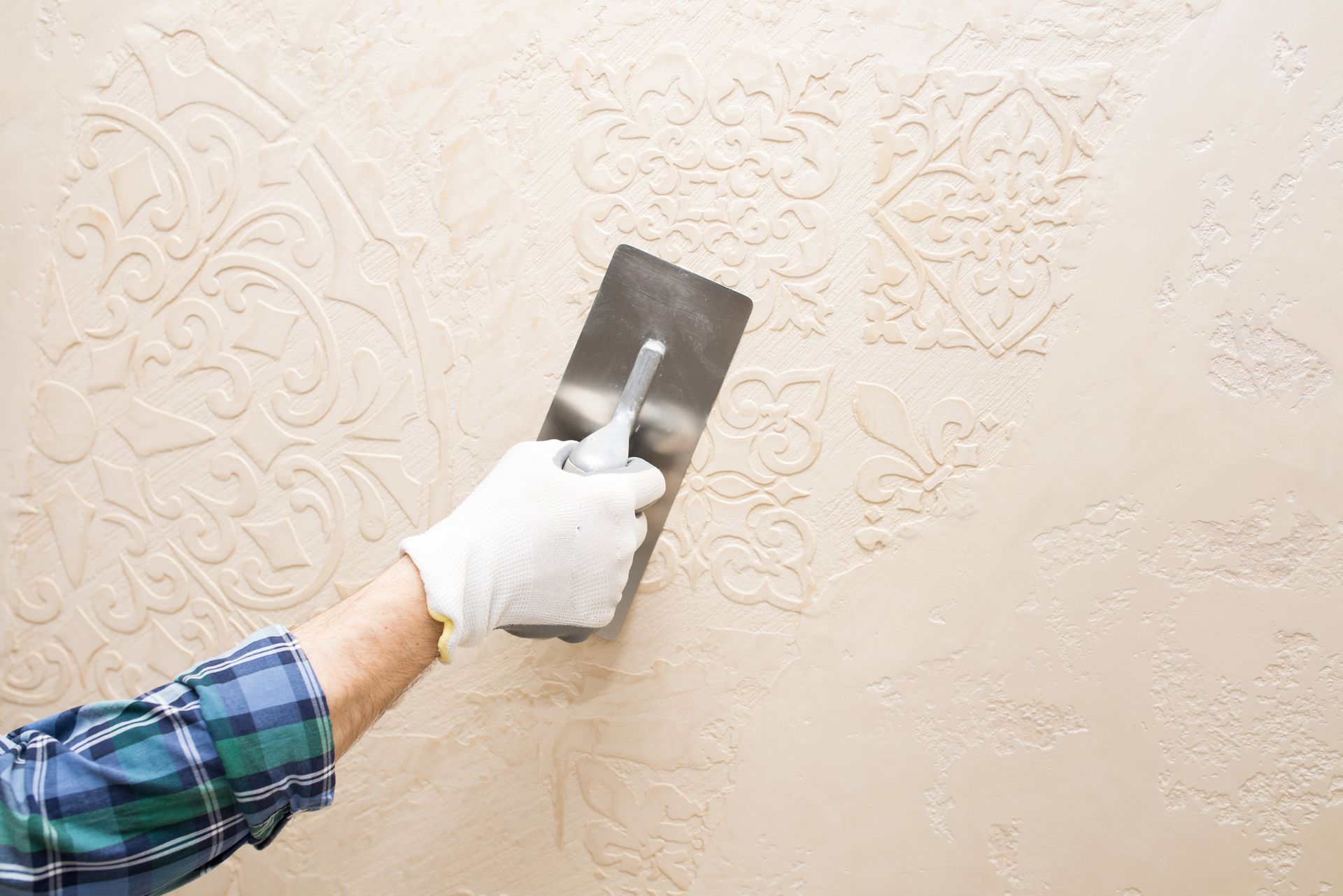 A person with a gloved hand uses a trowel to apply textured plaster to a wall with ornate patterns.