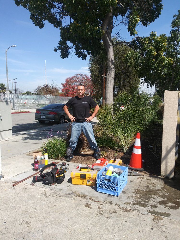 Man in black shirt and jeans stands near tools and a tree.