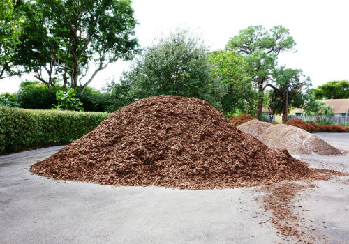 Large piles of brown mulch on a paved path beside green hedges and trees in a park