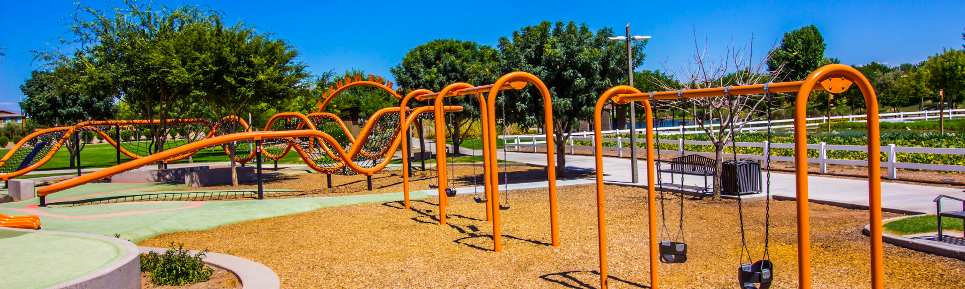 Orange playground fitness equipment in a sunny park with trees and a path nearby