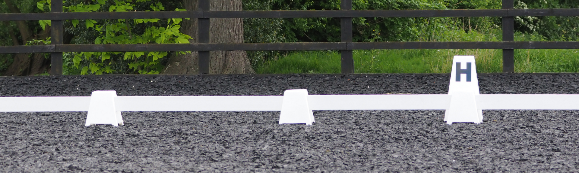 Dressage arena marker on a gravel track with white rail and fence, labeled “H” on the right