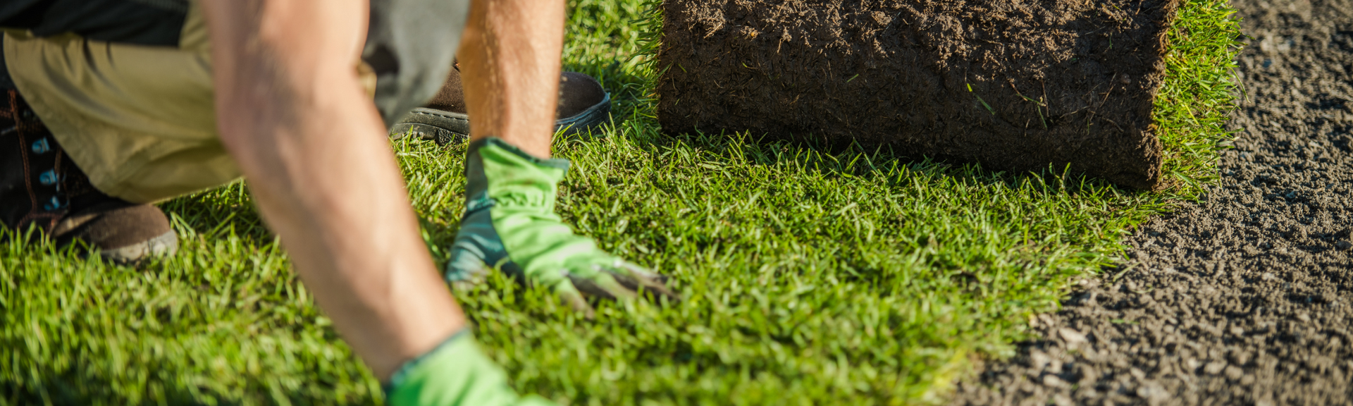 Person in green gloves digging soil beside a stone border in a grassy garden area