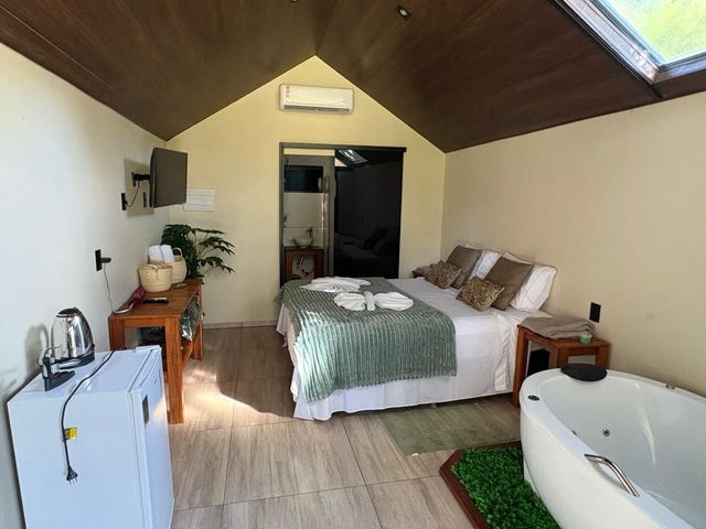 Quarto pequeno com cama, frigobar, penteadeira, vaso de planta e banheira sob um teto inclinado de madeira.