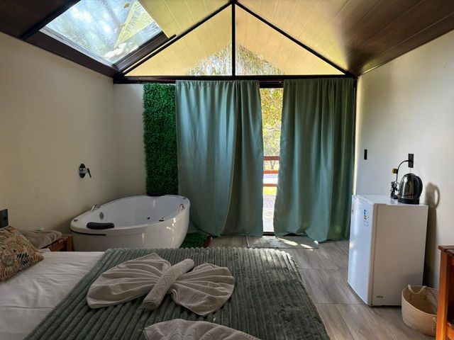 Quarto iluminado com banheira, cama, cortinas verdes e teto com claraboia.