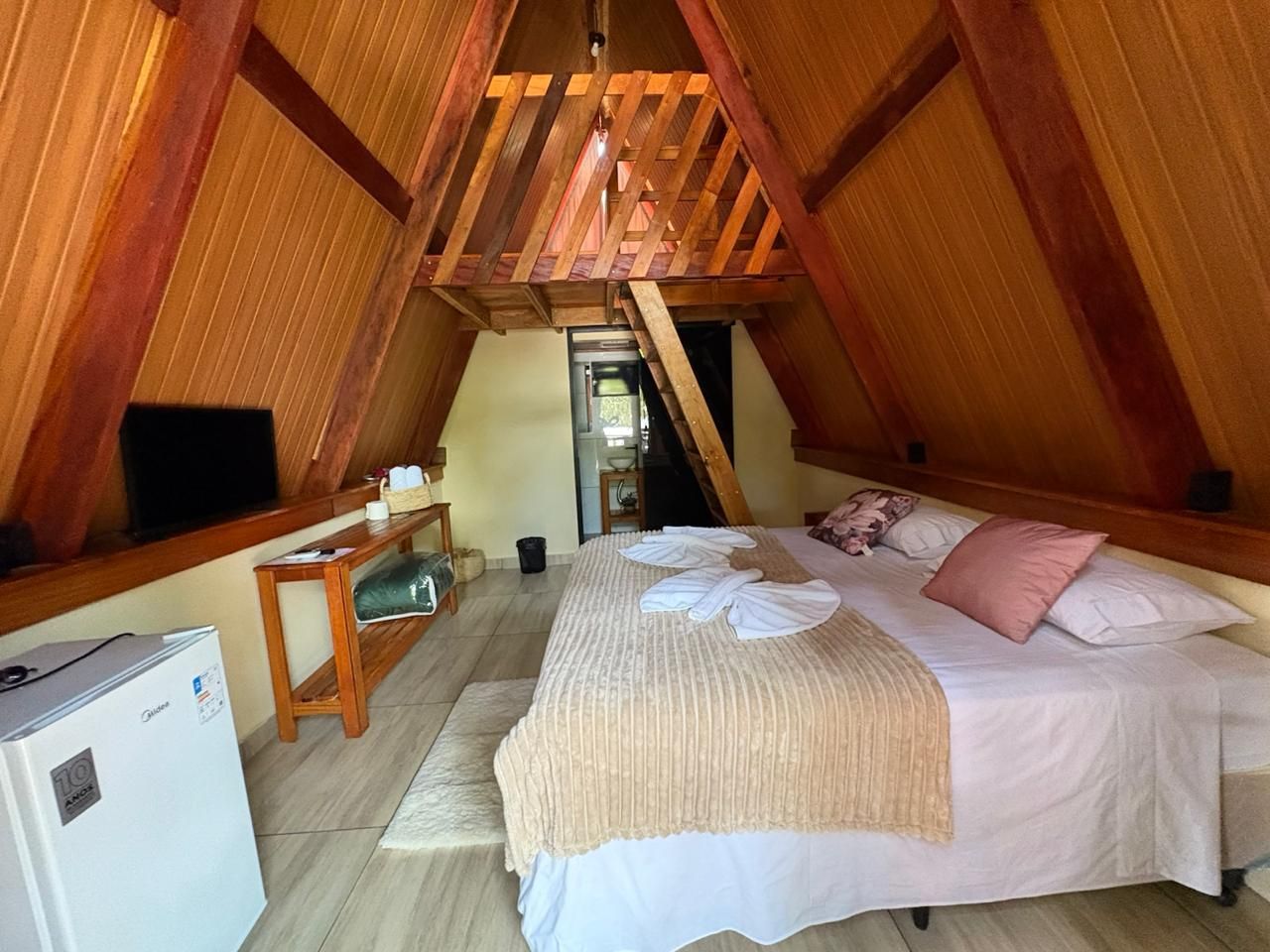 Quarto aconchegante em estilo loft com paredes revestidas de madeira, cama, escrivaninha pequena, TV e frigobar.