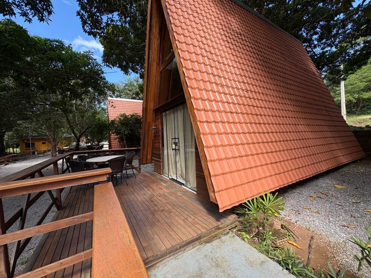 Cabana de madeira em forma de A com telhado vermelho íngreme ao lado de um deck e mesas de piquenique sob as árvores.