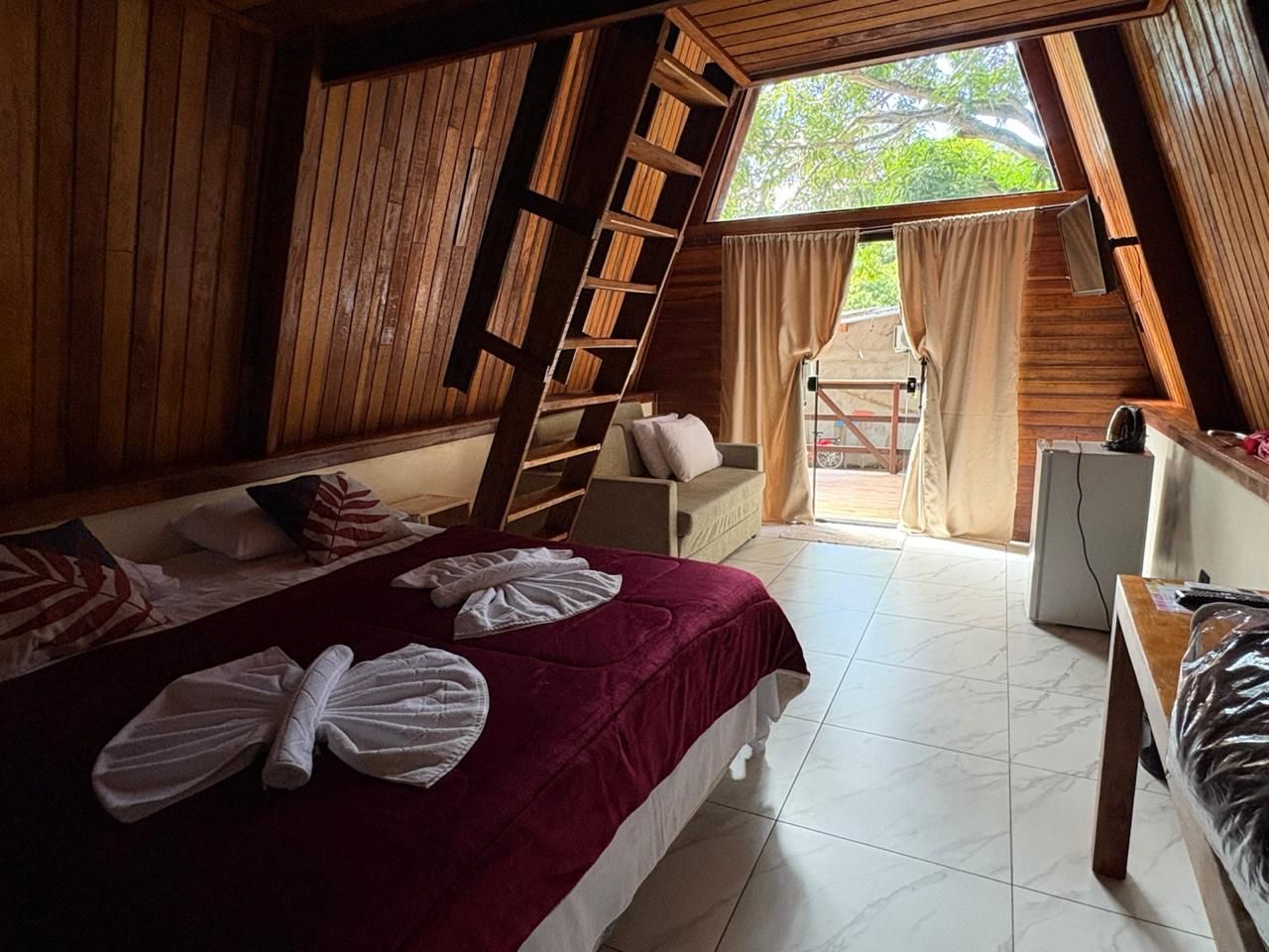 Um quarto aconchegante em uma cabana de madeira com uma colcha vermelha, toalhas dobradas e cortinas que se abrem para uma varanda.