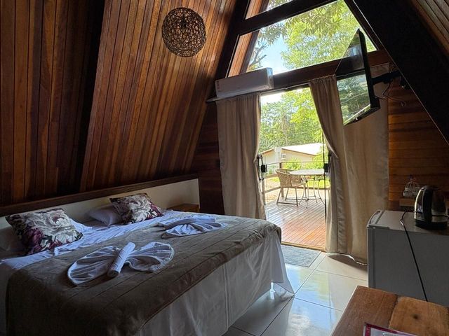 Quarto aconchegante com teto inclinado de madeira, cama desarrumada e cortinas abertas que dão para um pátio ensolarado.