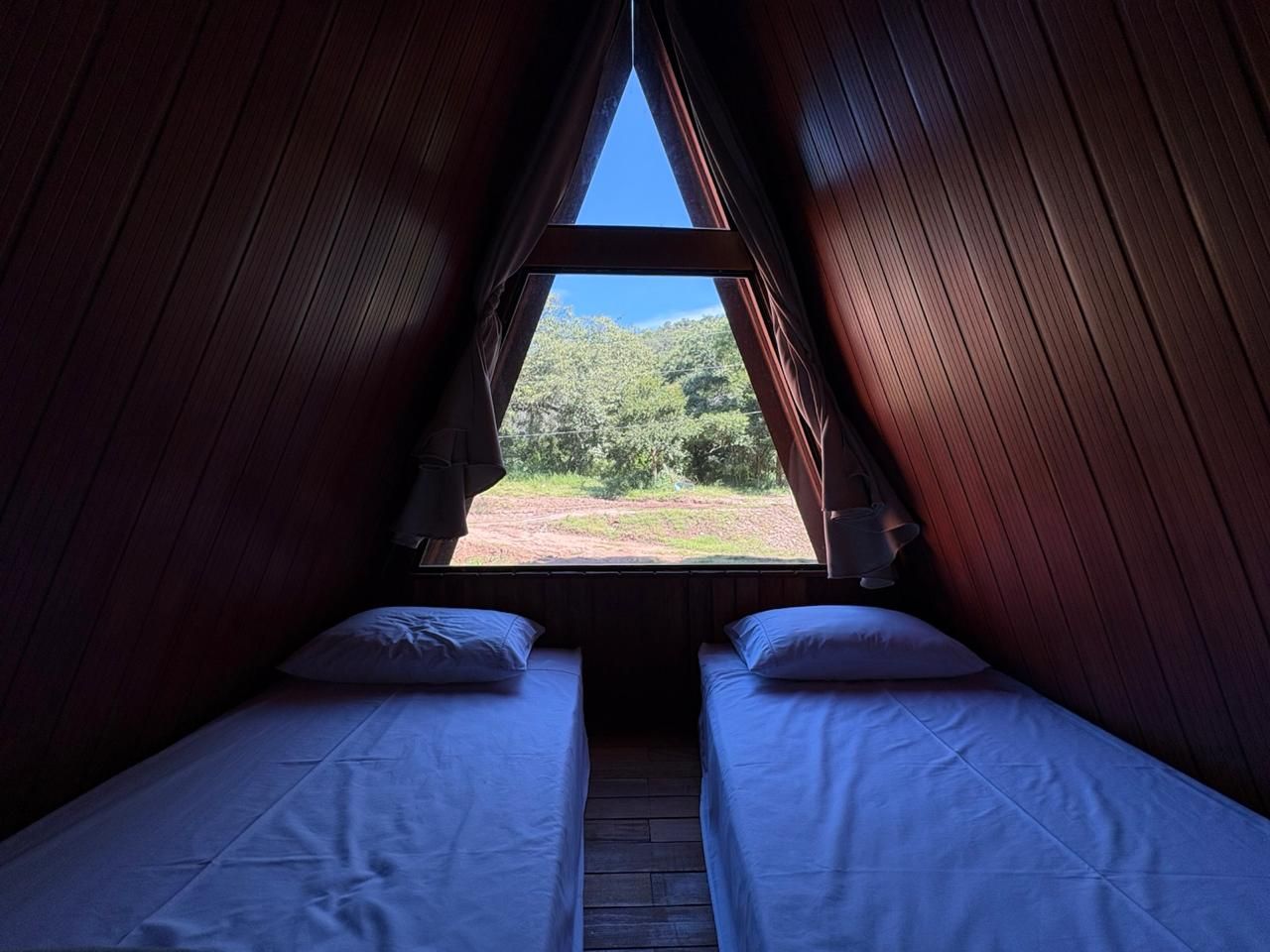 Duas camas de solteiro dentro de uma cabine triangular, de frente para uma grande janela com vista para as árvores.