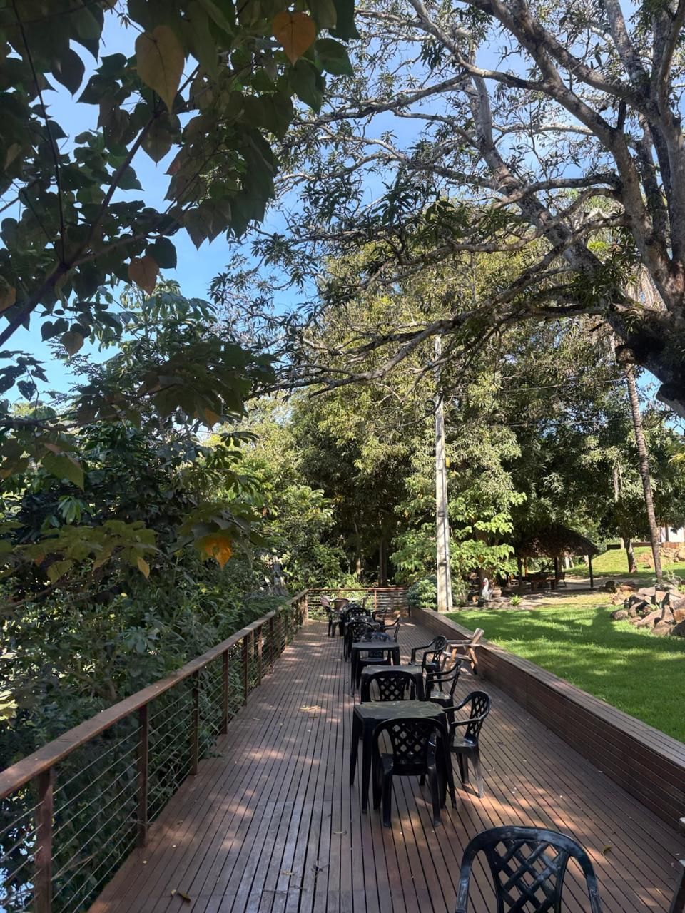 Caminho arborizado ao ar livre com mesas e cadeiras pretas em um deck de madeira sob a luz do sol filtrada pelas árvores.