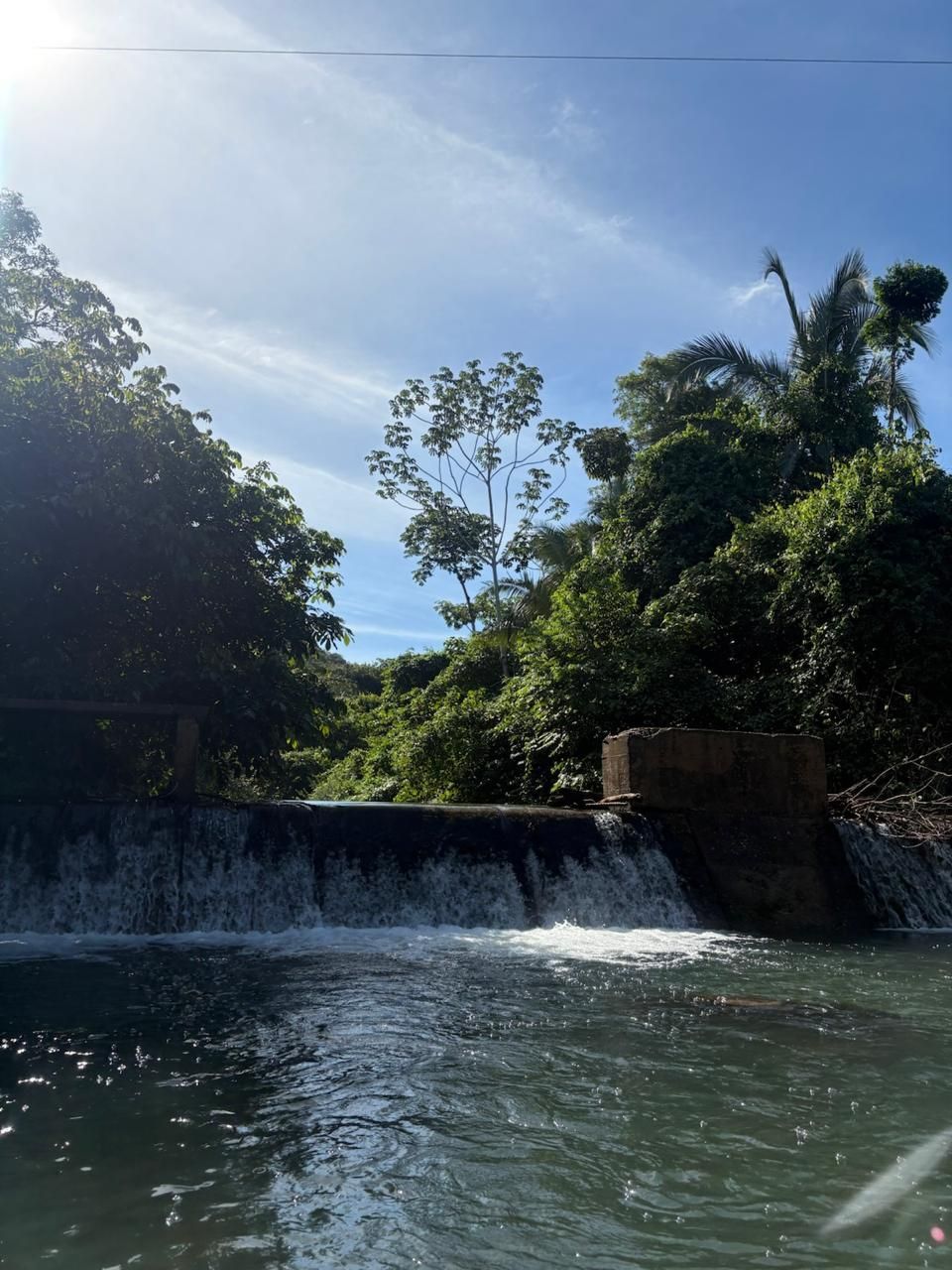 Cachoeira iluminada pelo sol desaguando em uma piscina tranquila, cercada por árvores verdejantes sob um céu azul límpido.