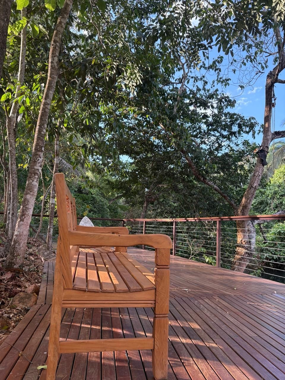 Banco de madeira em um deck ensolarado entre árvores, com vista para uma encosta arborizada sob um céu azul.