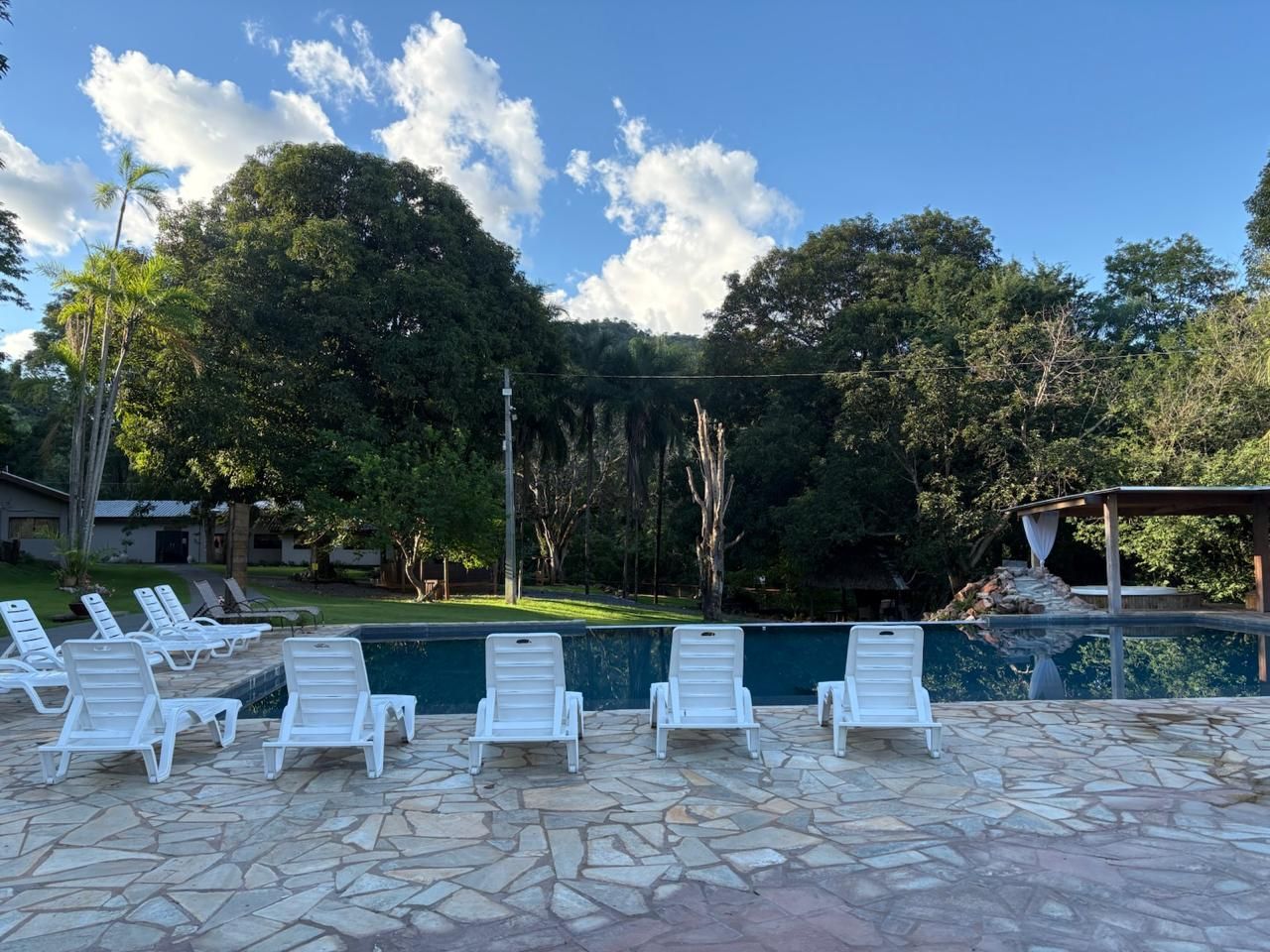 Pátio à beira da piscina com espreguiçadeiras brancas, piscina azul e árvores exuberantes sob um céu claro.