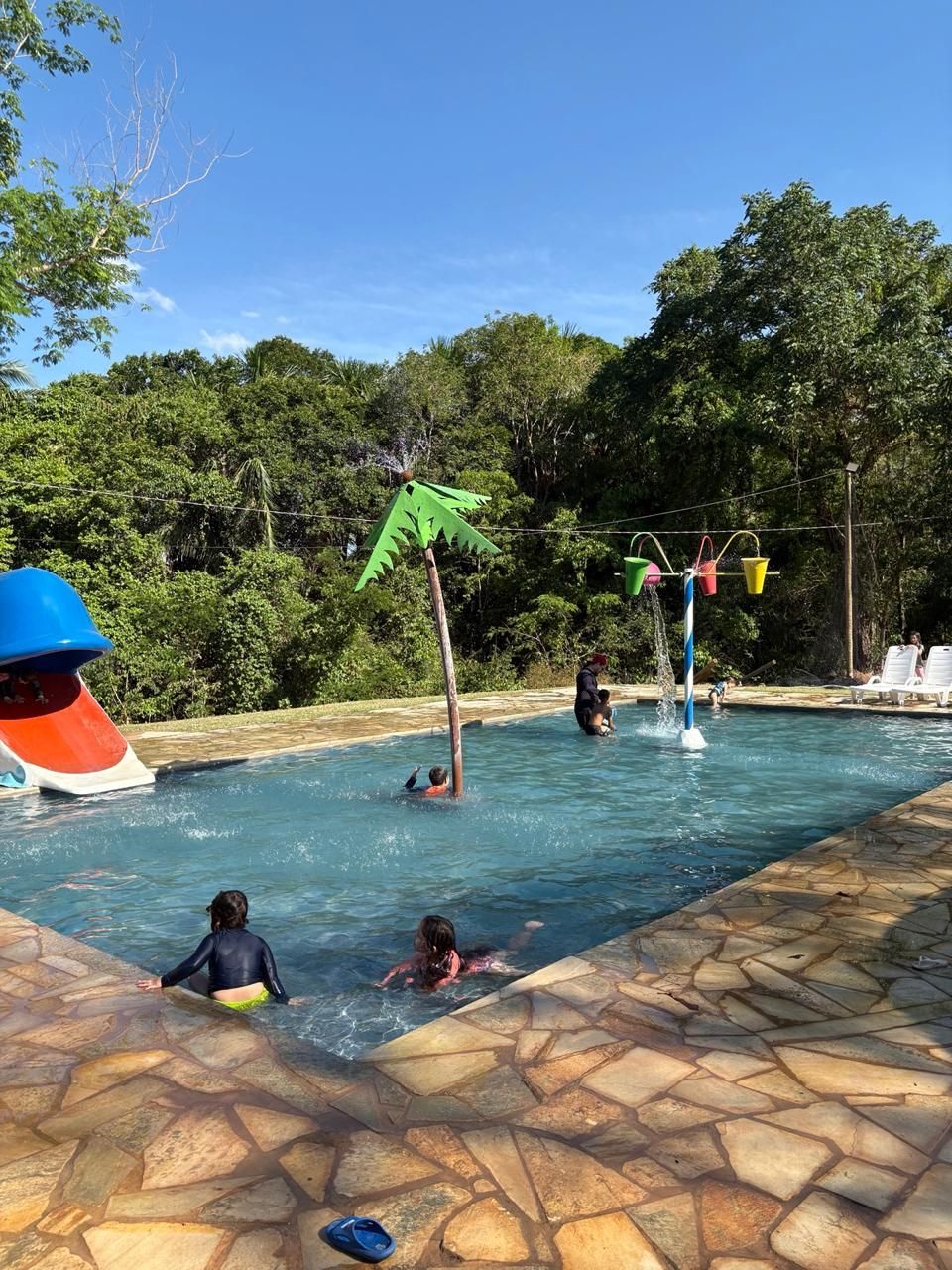 Crianças brincam em uma piscina no quintal com um escorregador verde, brinquedos aquáticos e deck de pedra sob um céu azul.