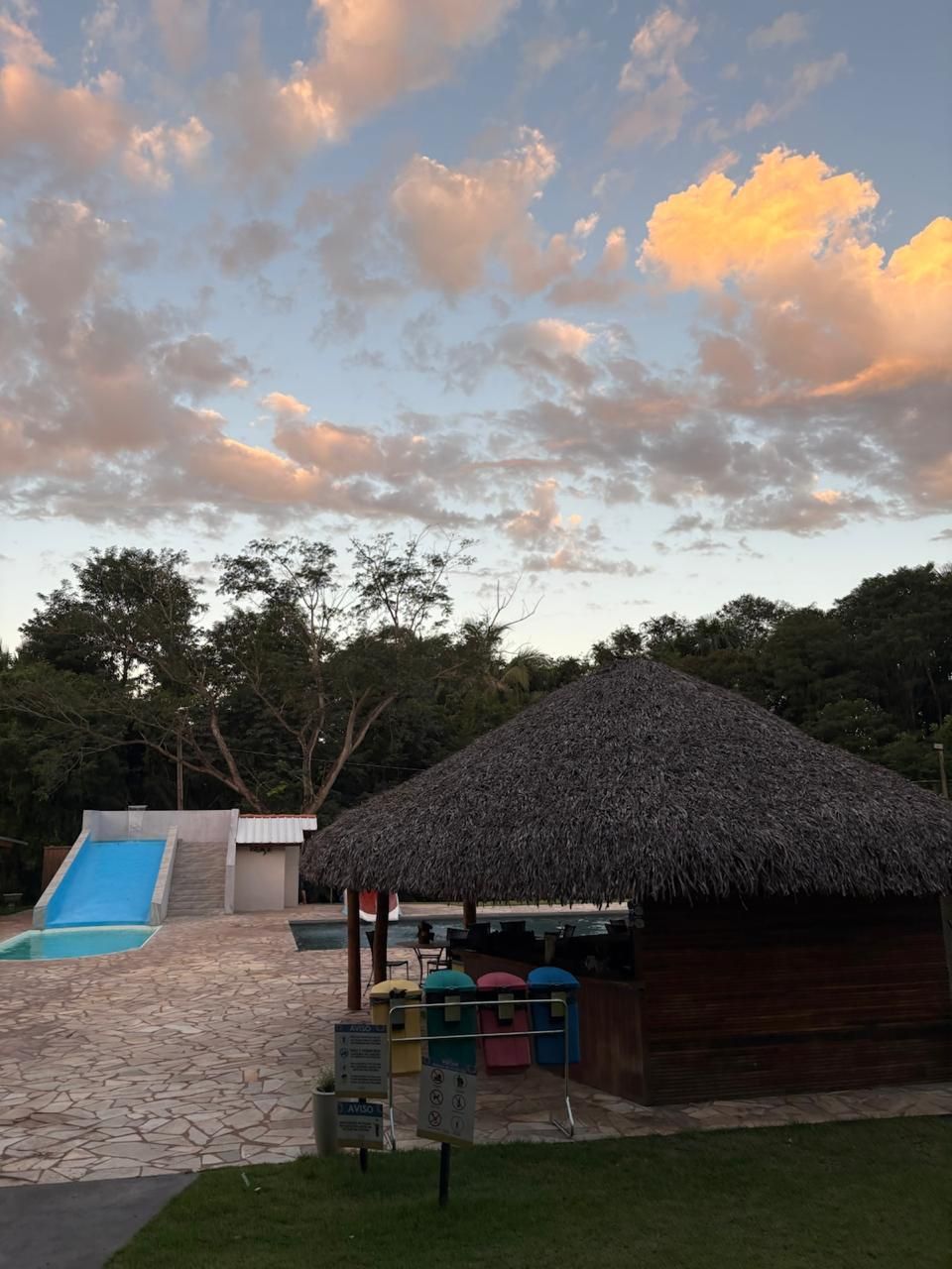Área da piscina do resort com bar de teto de palha, tobogã azul e pôr do sol sobre as árvores.