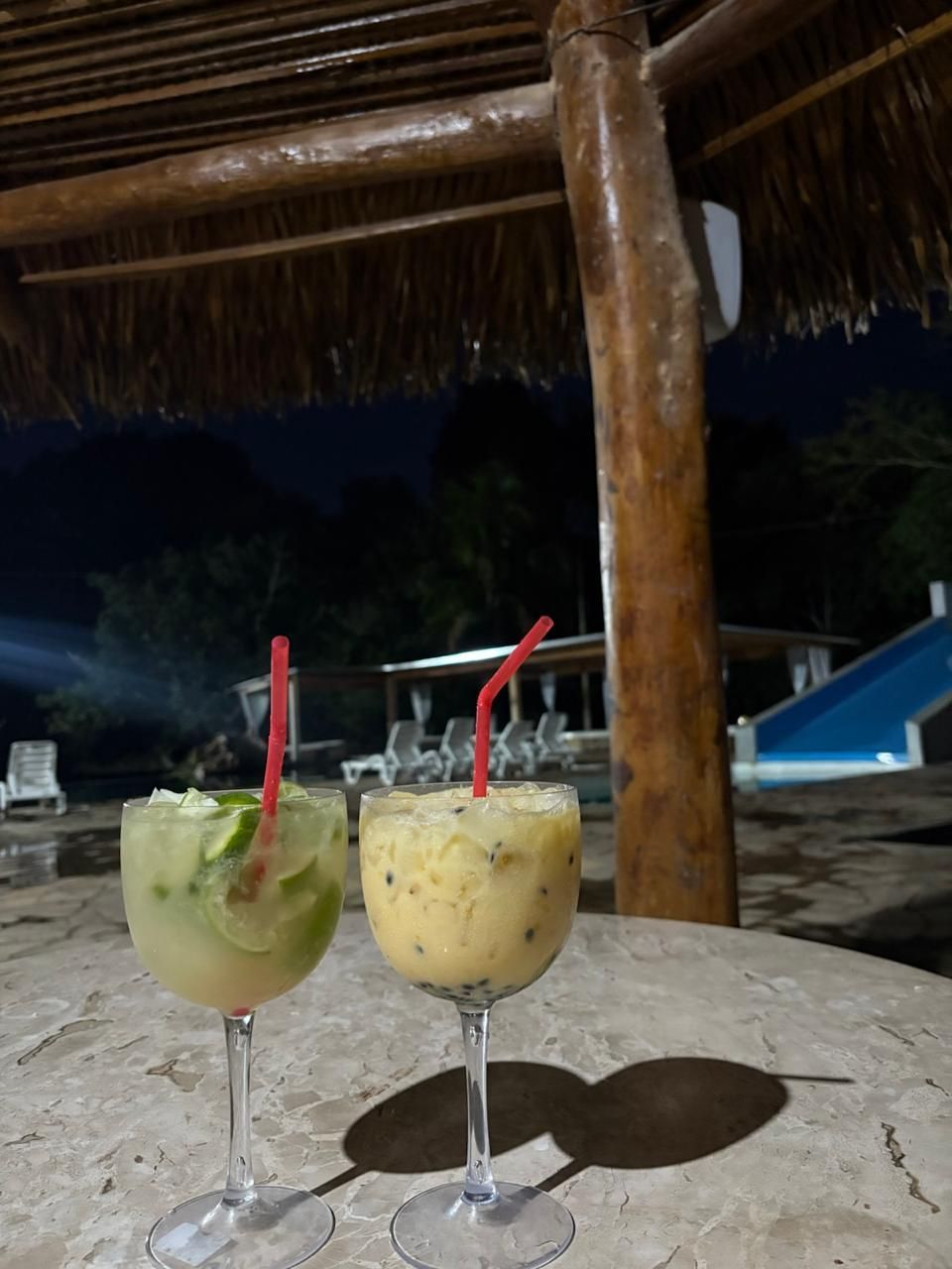 Dois coquetéis com canudos vermelhos sobre uma mesa sob um telhado de palha à noite.
