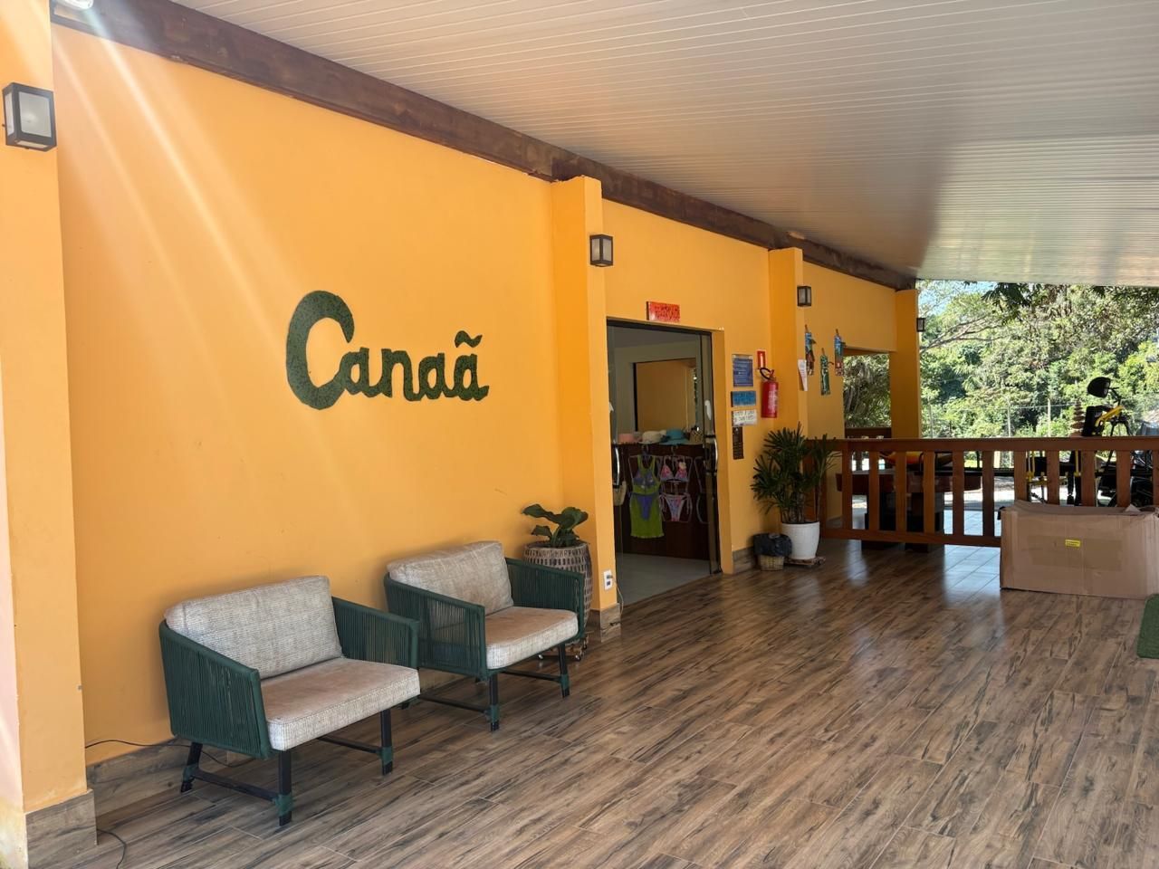 Varanda coberta de cor laranja com placa “Canã”, duas cadeiras e corrimão de madeira com vista para a vegetação.