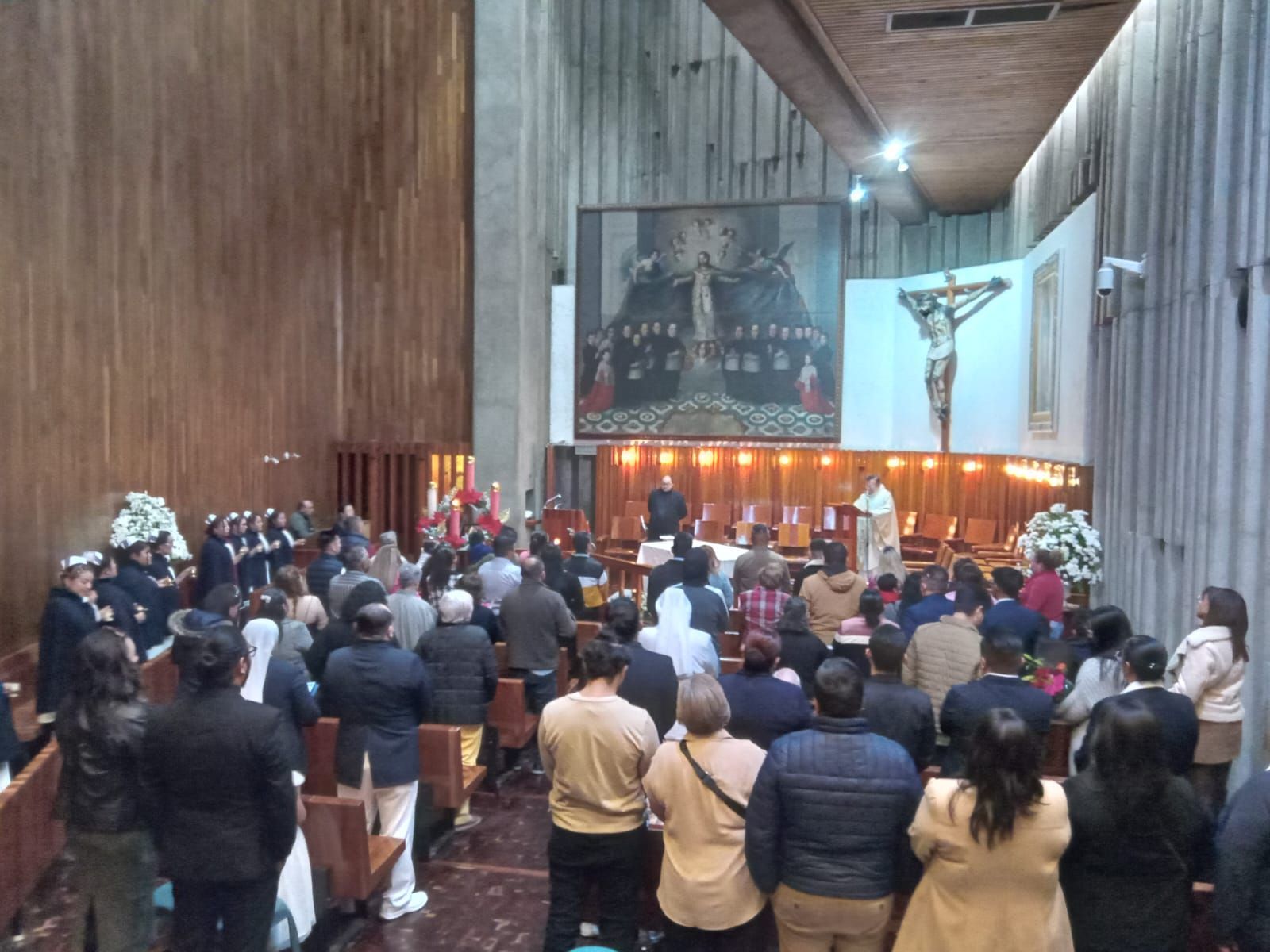 Personas en un servicio religioso, frente a un crucifijo, con sacerdotes y una pintura de figuras.