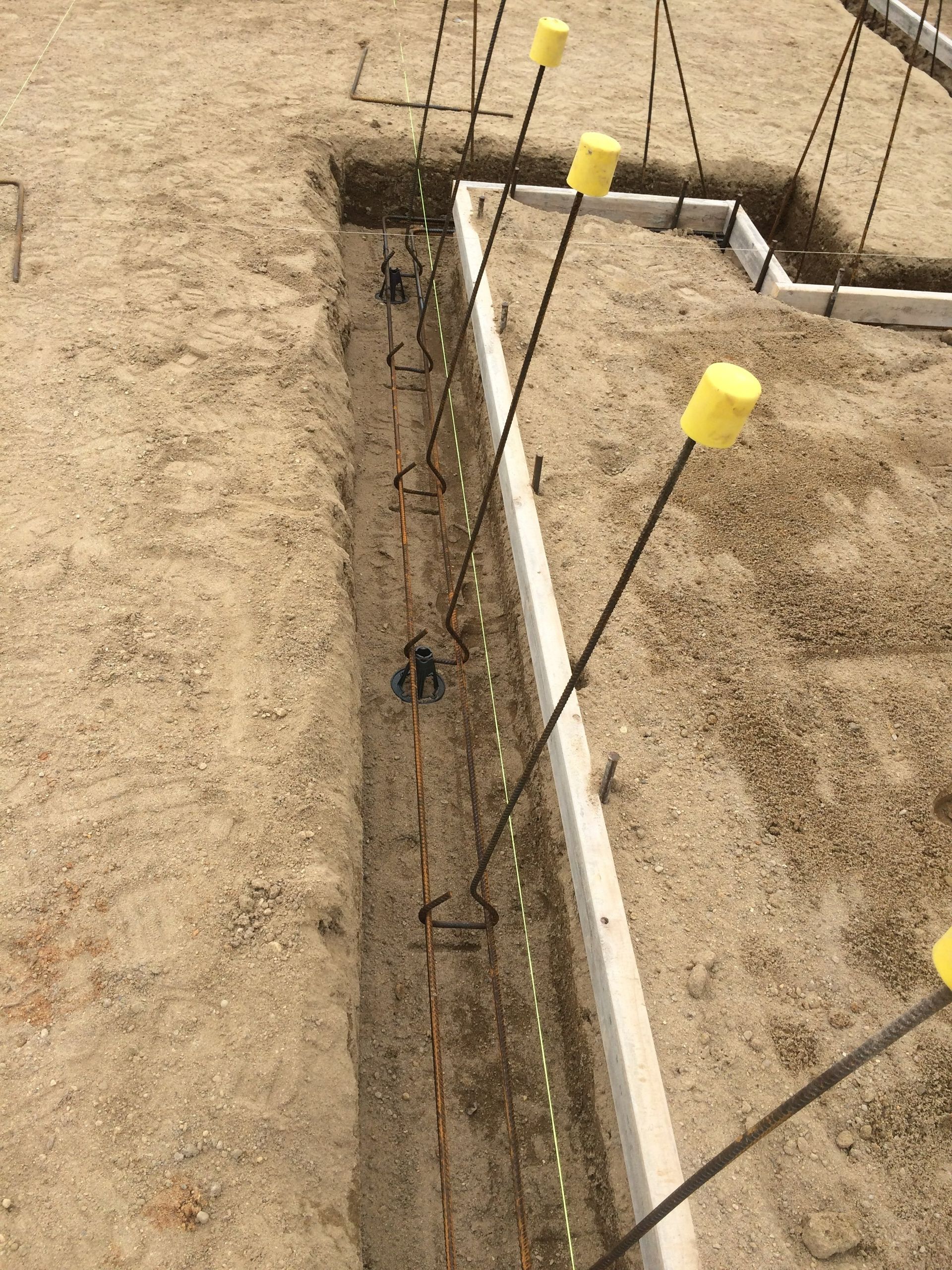 Construction site: Trench with rebar and yellow capped stakes, next to a concrete form.