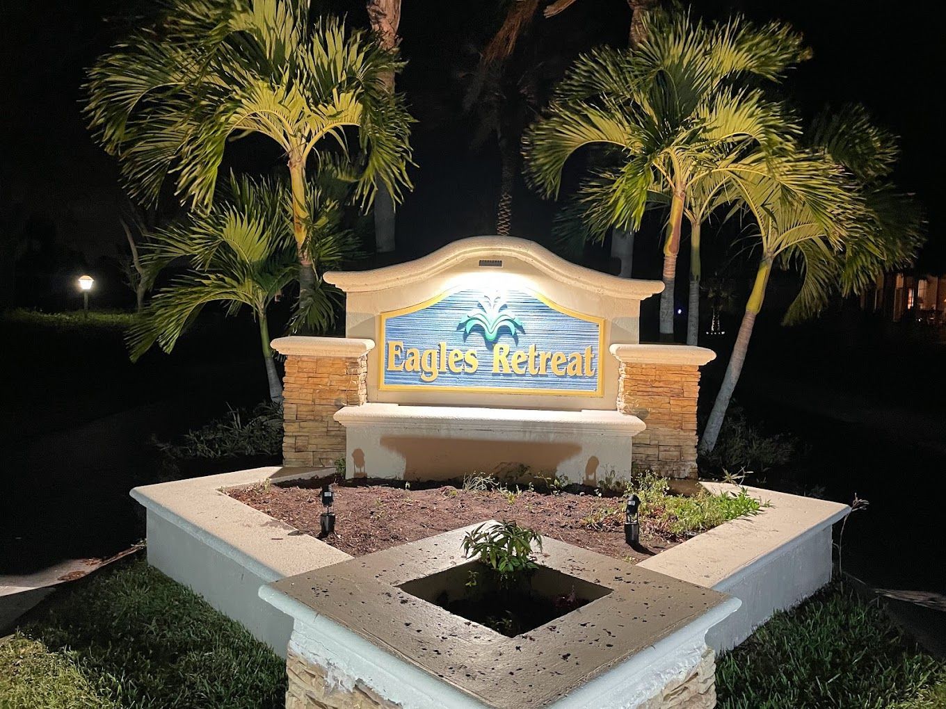 Lighted entrance sign for Eagles Retreat at night, featuring a stone-colored monument, palm trees, and a small planter.