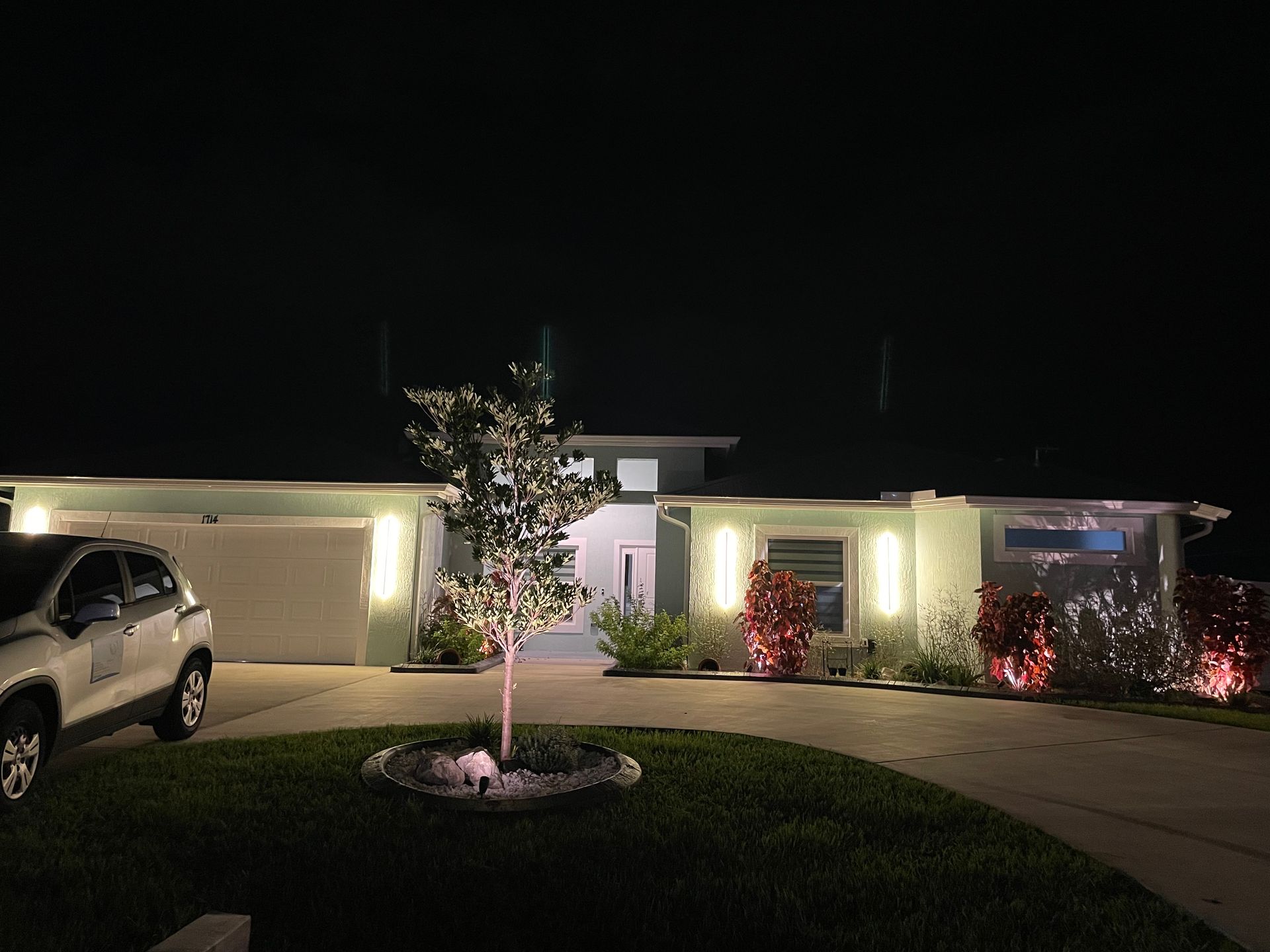 Night view of a residential home with illuminated uplighting on a tree and the house exterior, featuring a parked SUV.