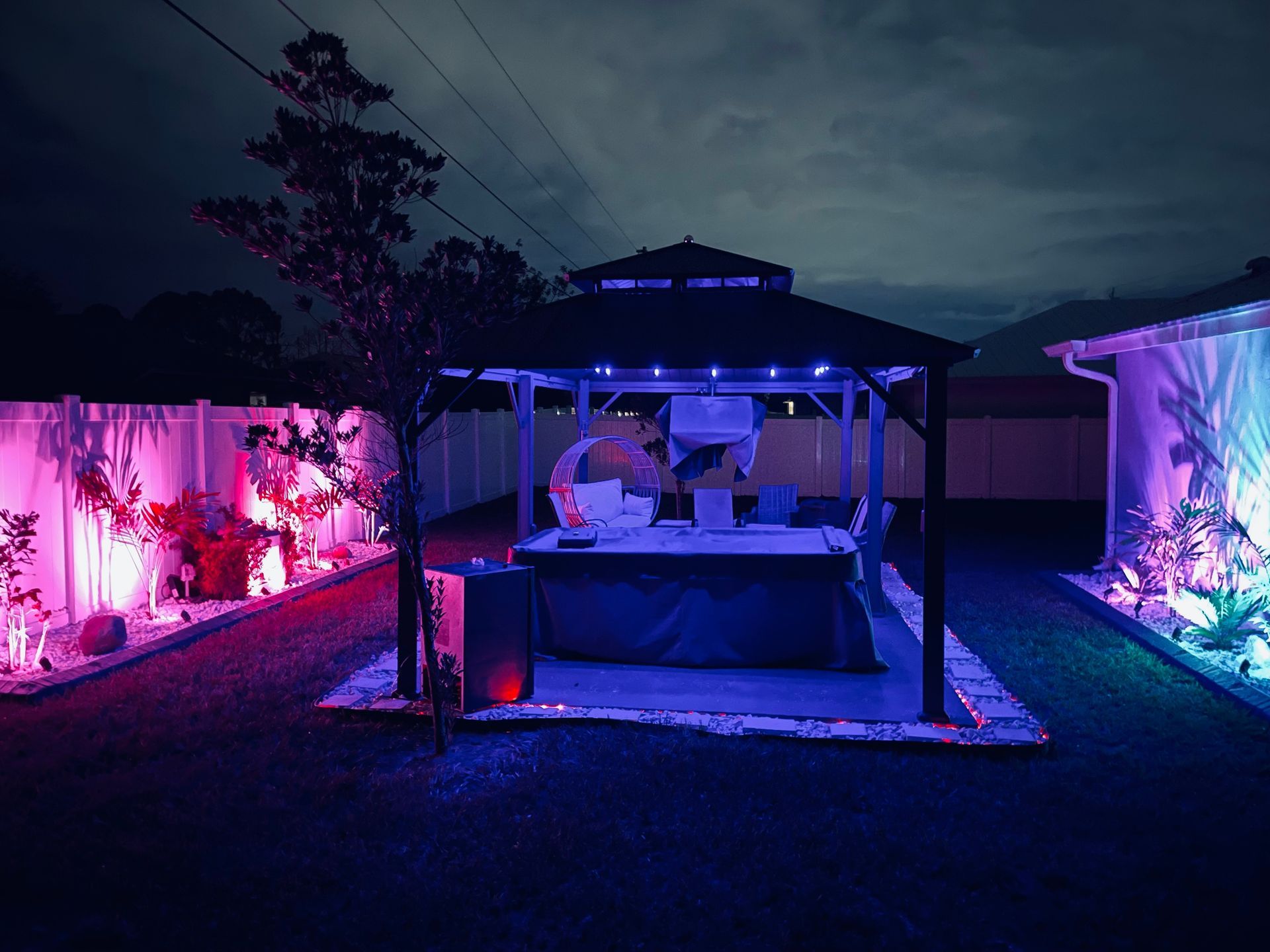 A gazebo with a hot tub is illuminated by blue lights in a backyard at night, flanked by purple-lit landscaping.